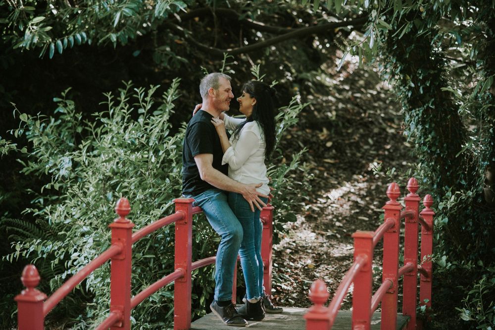 A couple sharing a romantic moment on a red bridge surrounded by green foliage.