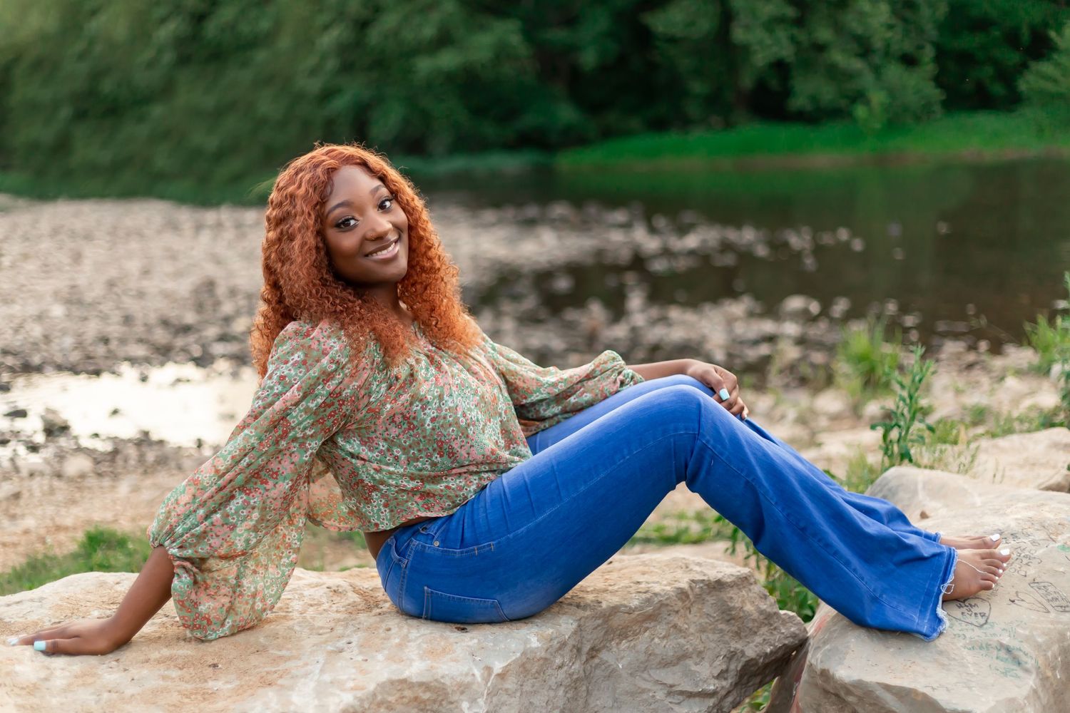 Woman with vibrant red hair poses on a rock by a river wearing blue jeans and a patterned top.