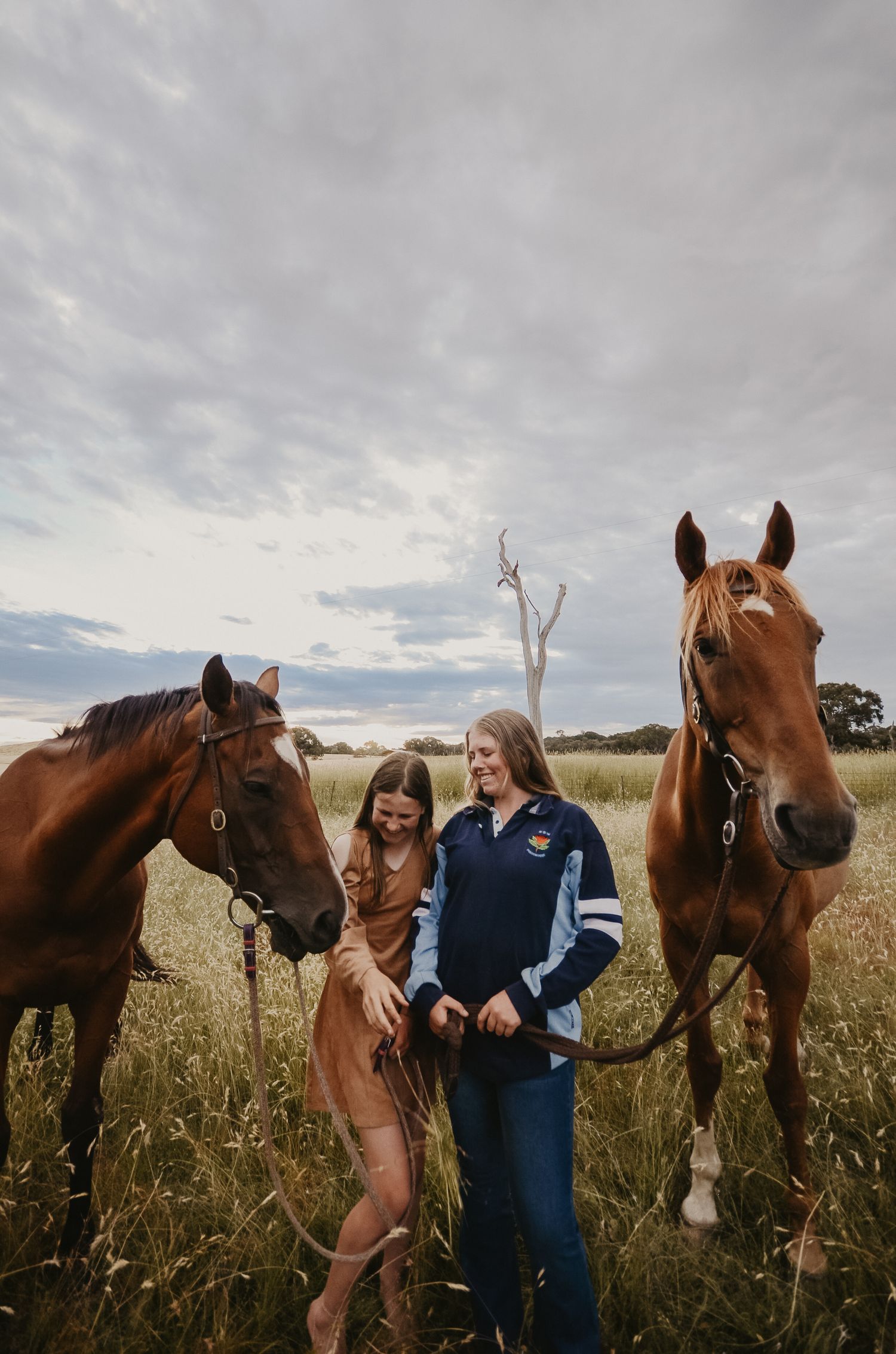 two girls laughing with each other in a paddock with long grass and their two horses standing beside them