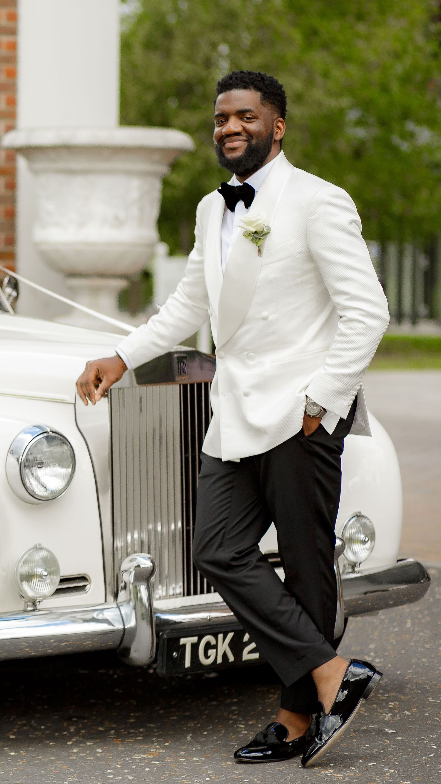 Stylish groom in white tuxedo jacket posing beside vintage Rolls Royce.