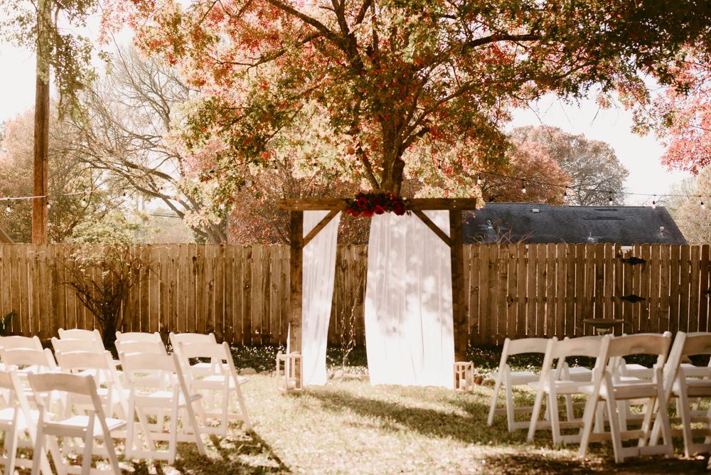 A rustic outdoor wedding ceremony setup with white chairs and an arch framed by autumn trees.