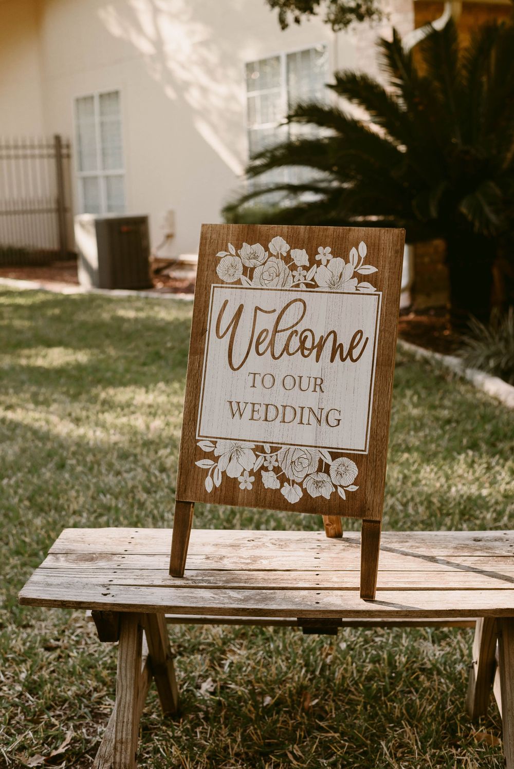 Rustic wooden welcome sign with floral design displayed on bench at outdoor wedding venue.