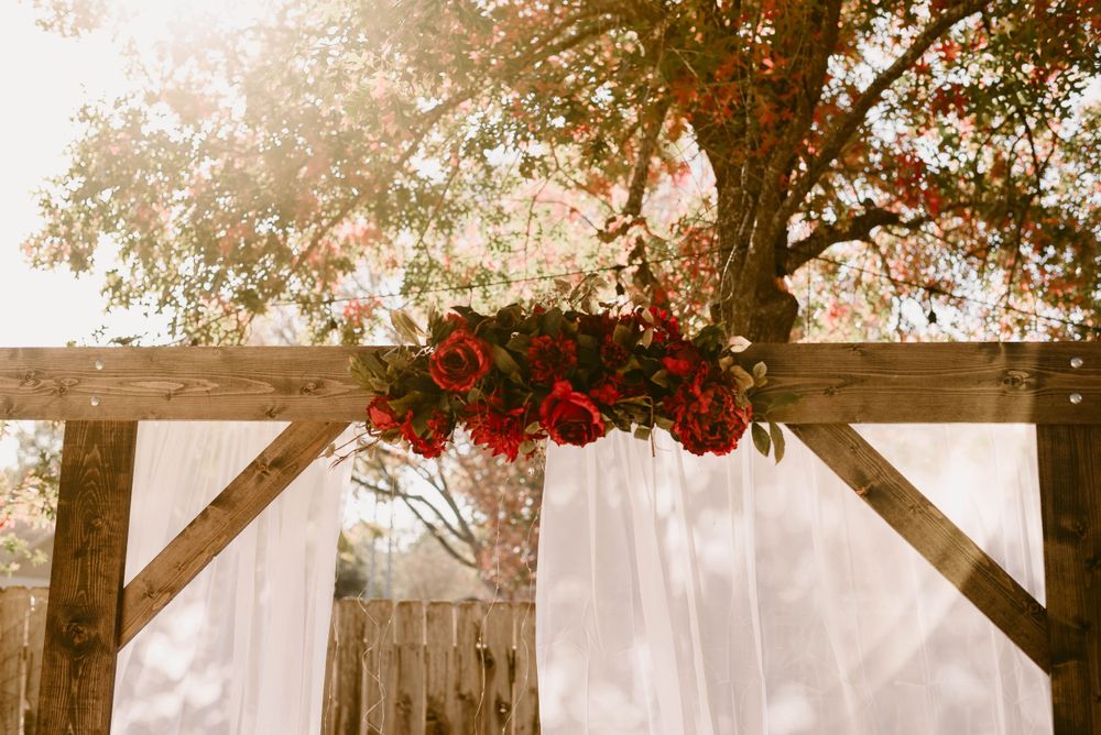 Wooden wedding arch decorated with red roses and white fabric against sunlit trees.