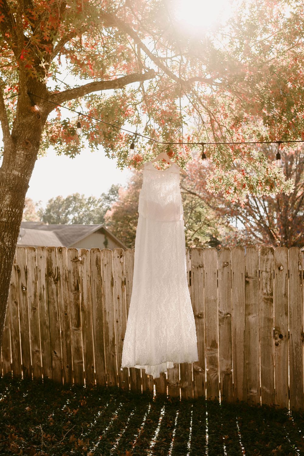 A white lace wedding dress hangs from a tree branch against a wooden fence in warm sunlight.