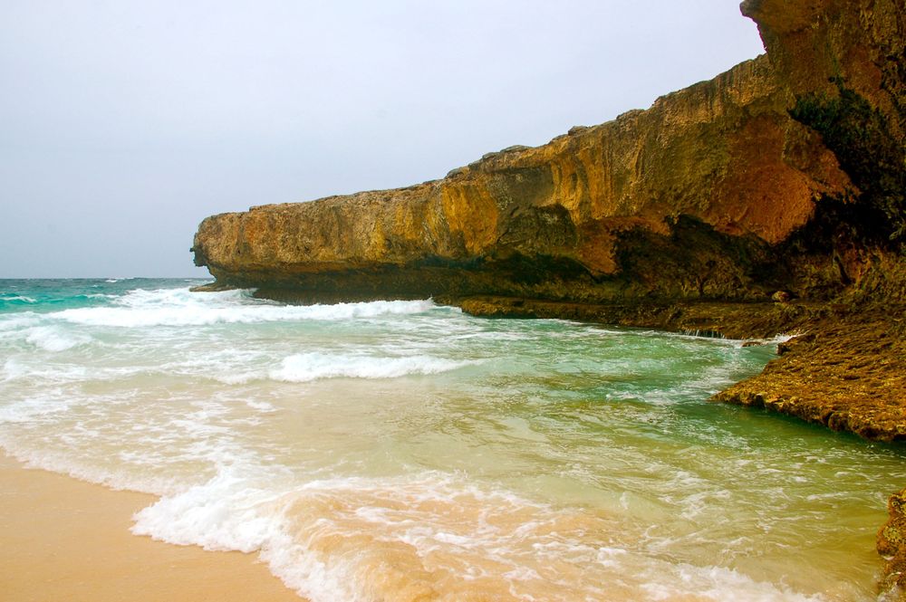 Rugged limestone cliffs tower over a sandy beach as waves roll into shore.