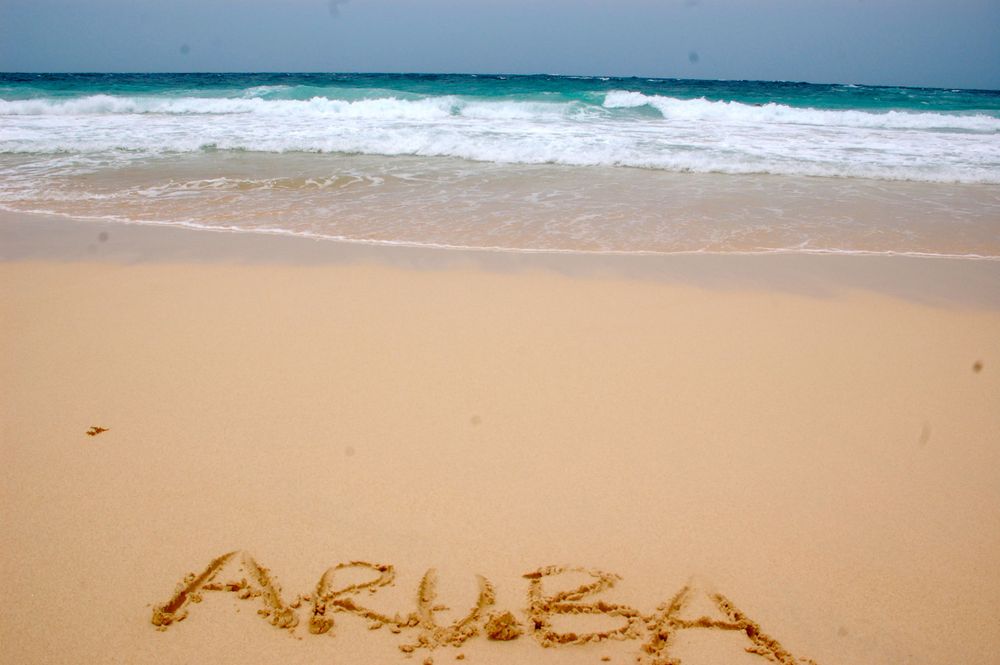 The word 'Aruba' written in sand on a pristine Caribbean beach with azure waters in the background.