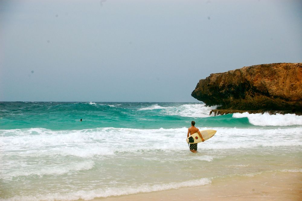 Surfer walks along a scenic beach with dramatic rocky cliffs and turquoise waters.
