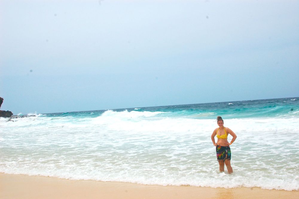 Beachgoer stands at the edge of crystal clear turquoise waters near rocky cliffs.