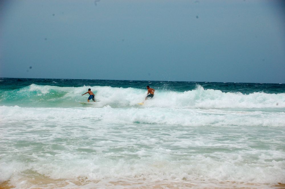 Two surfers ride turquoise waves in the distance on a bright tropical day.