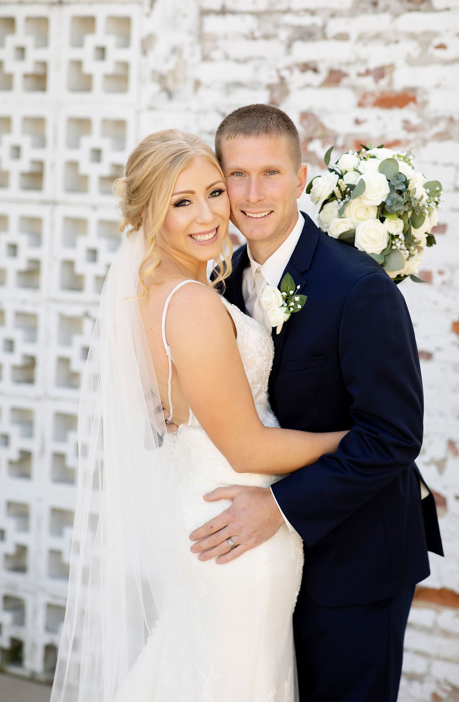 A joyful wedding portrait featuring a couple with white flowers against a textured wall background.