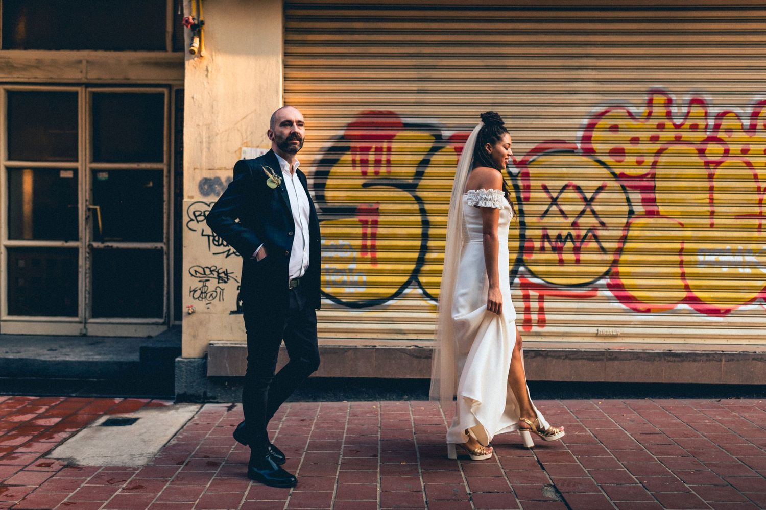 A wedding couple walks past colorful graffiti on an urban street.