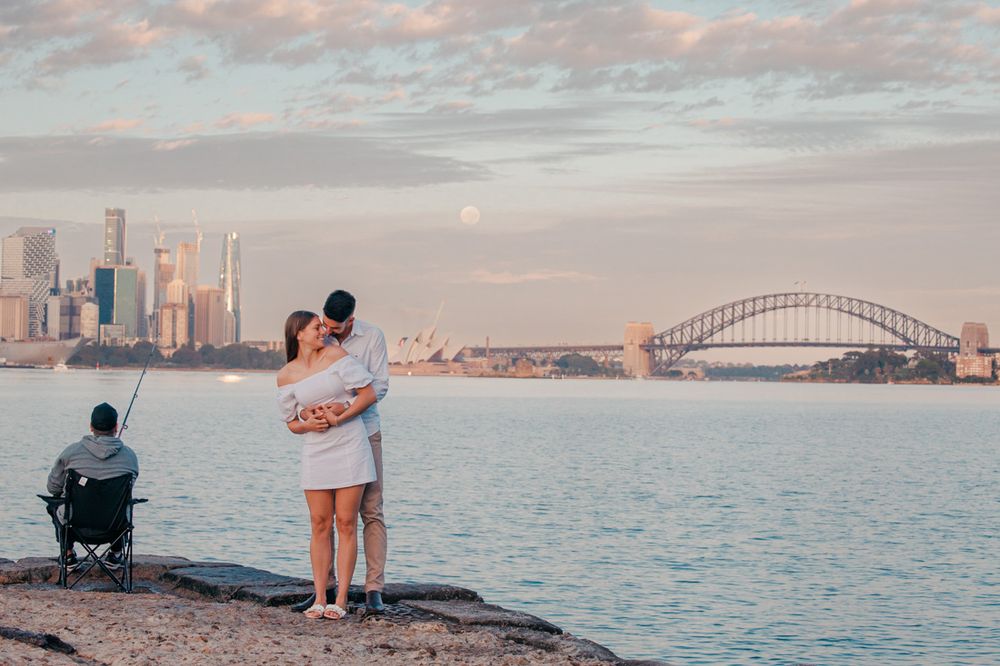 Romantic couple embracing at sunset with Sydney Harbour Bridge and city skyline in the background.