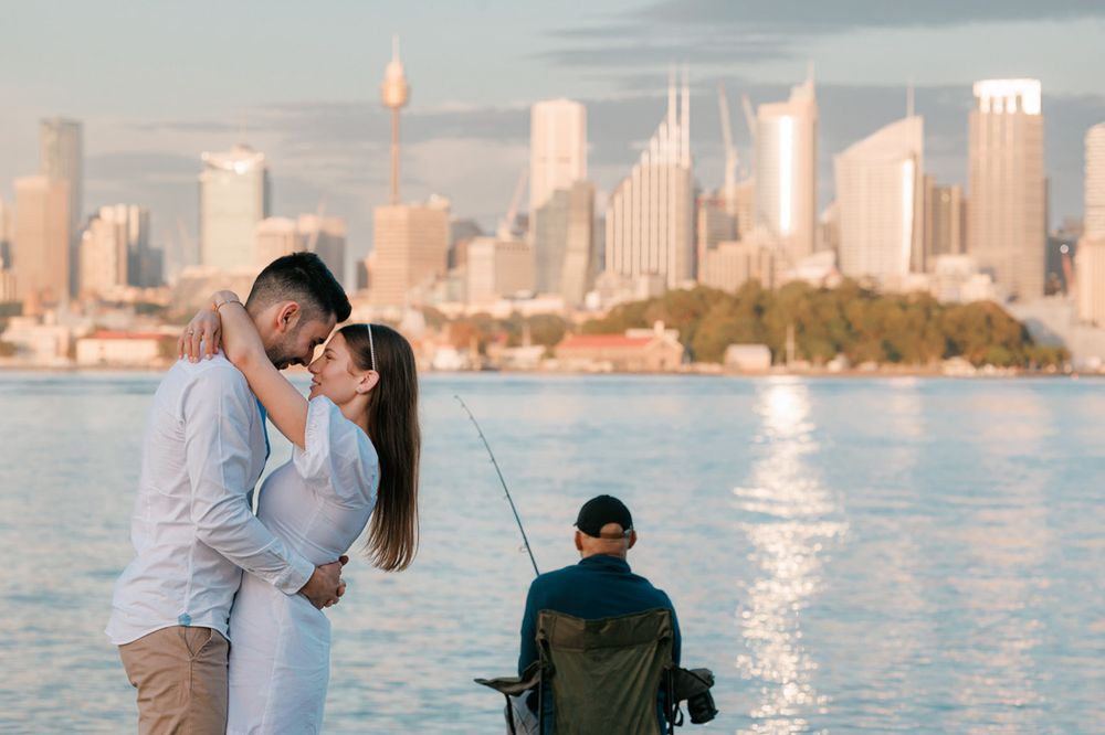 Waterfront scene with fisherman and couple against Sydney city backdrop.