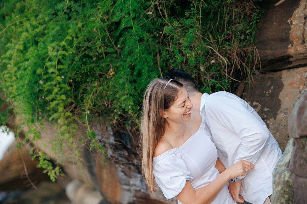 Romantic couple embraces near stone wall surrounded by lush greenery during outdoor photoshoot.