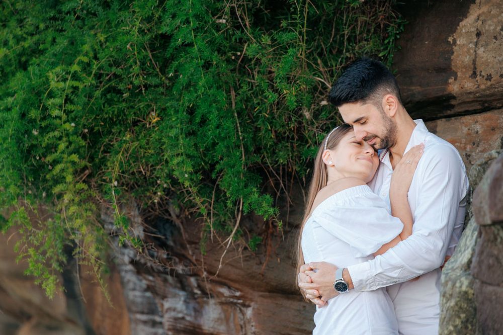 Couple shares intimate moment against rustic stone wall with green foliage overhead.