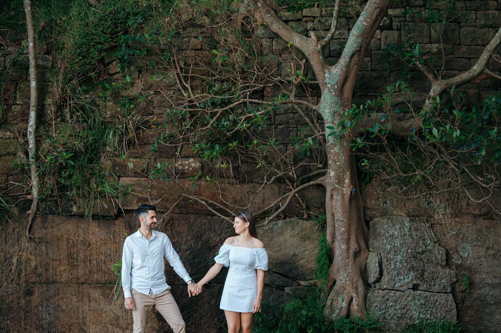 Couple poses together along stone wall path with trees creating natural archway.