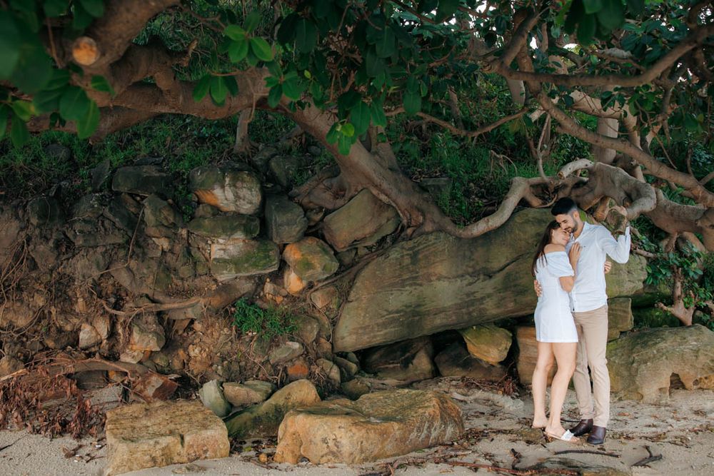 Couple explores rocky shoreline with trees and stone wall backdrop.