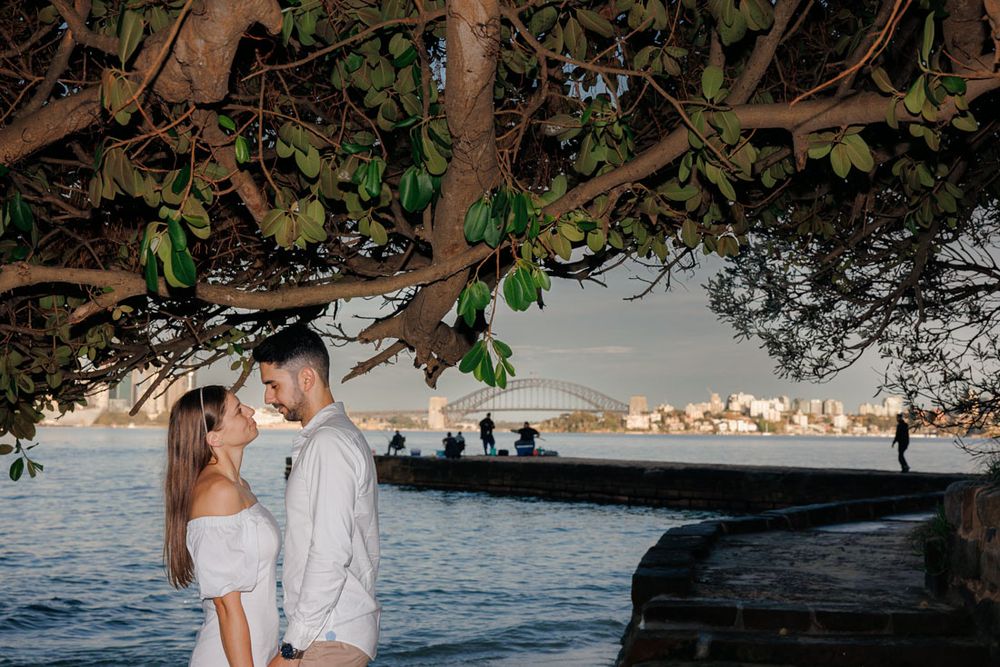 Couple shares romantic moment by water under overhanging tree branches.