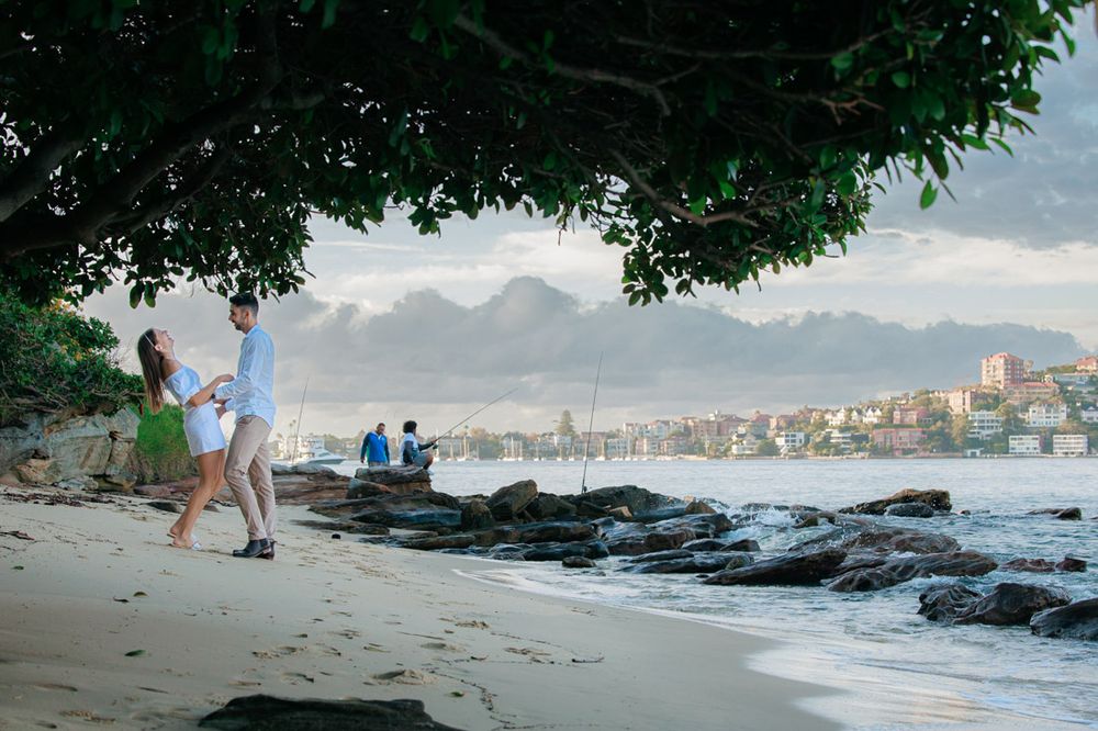 Rocky beach shoreline with lush green trees and city skyline in the background.
