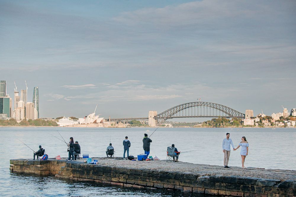 People standing on a long pier with Sydney Harbour Bridge and Opera House in the background.