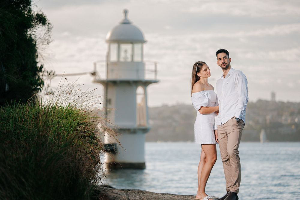 A romantic couple stands near a lighthouse overlooking a harbor during sunset.