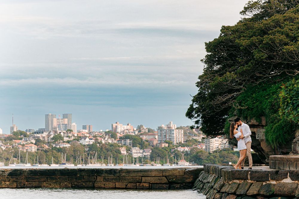 A couple embraces on a coastal path with a city skyline visible across the water.