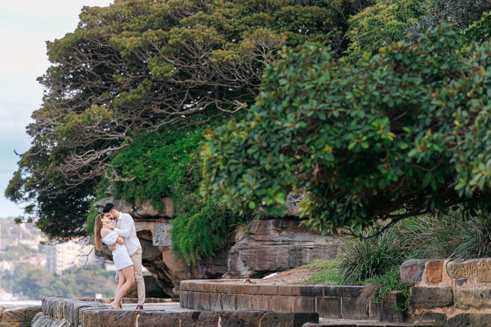 A couple shares an intimate moment on stone steps with a lighthouse in the background.