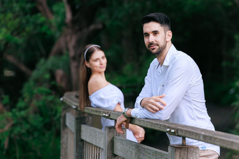 A couple leans against a wooden railing in a lush green park.