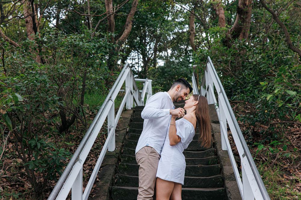 A couple embraces on a white bridge surrounded by trees.