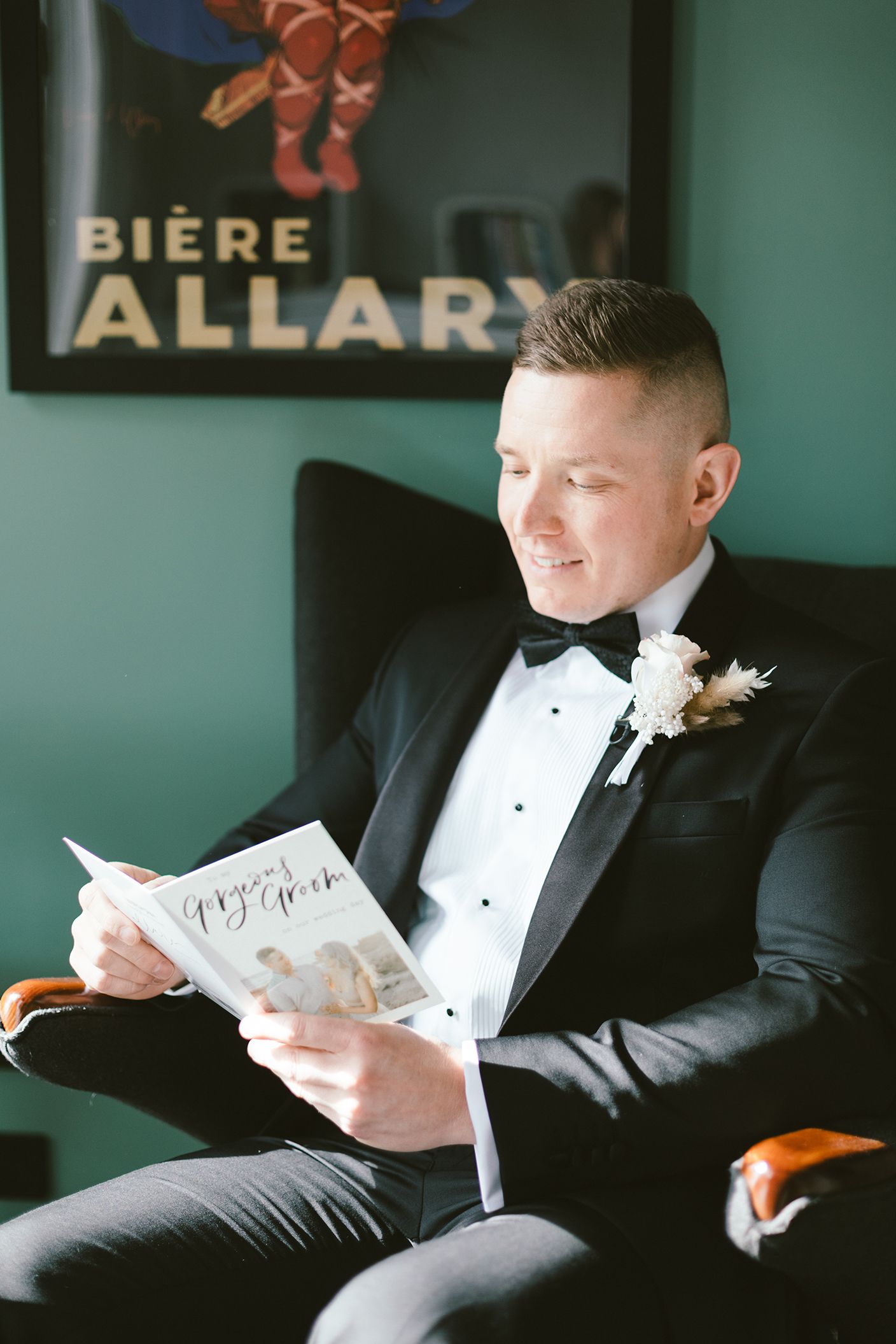 A groom in a tuxedo reads a document while sitting in a chair with a vintage Biere Allary poster in the background.