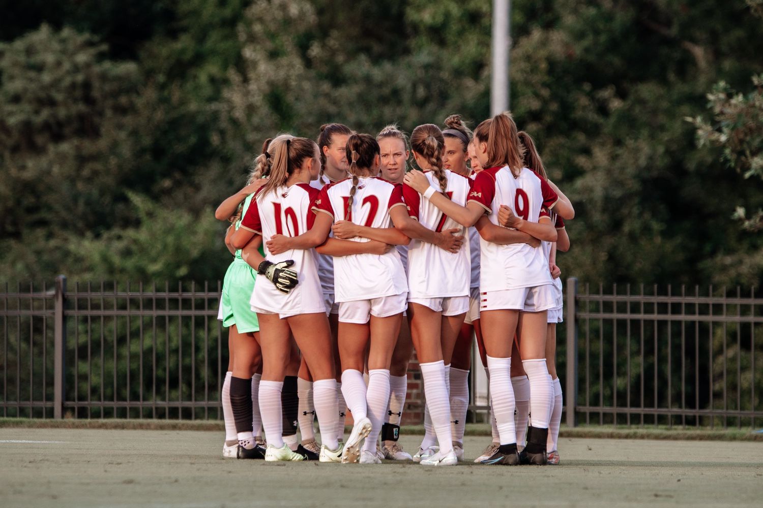 A women's soccer team huddled together on the field wearing white uniforms.