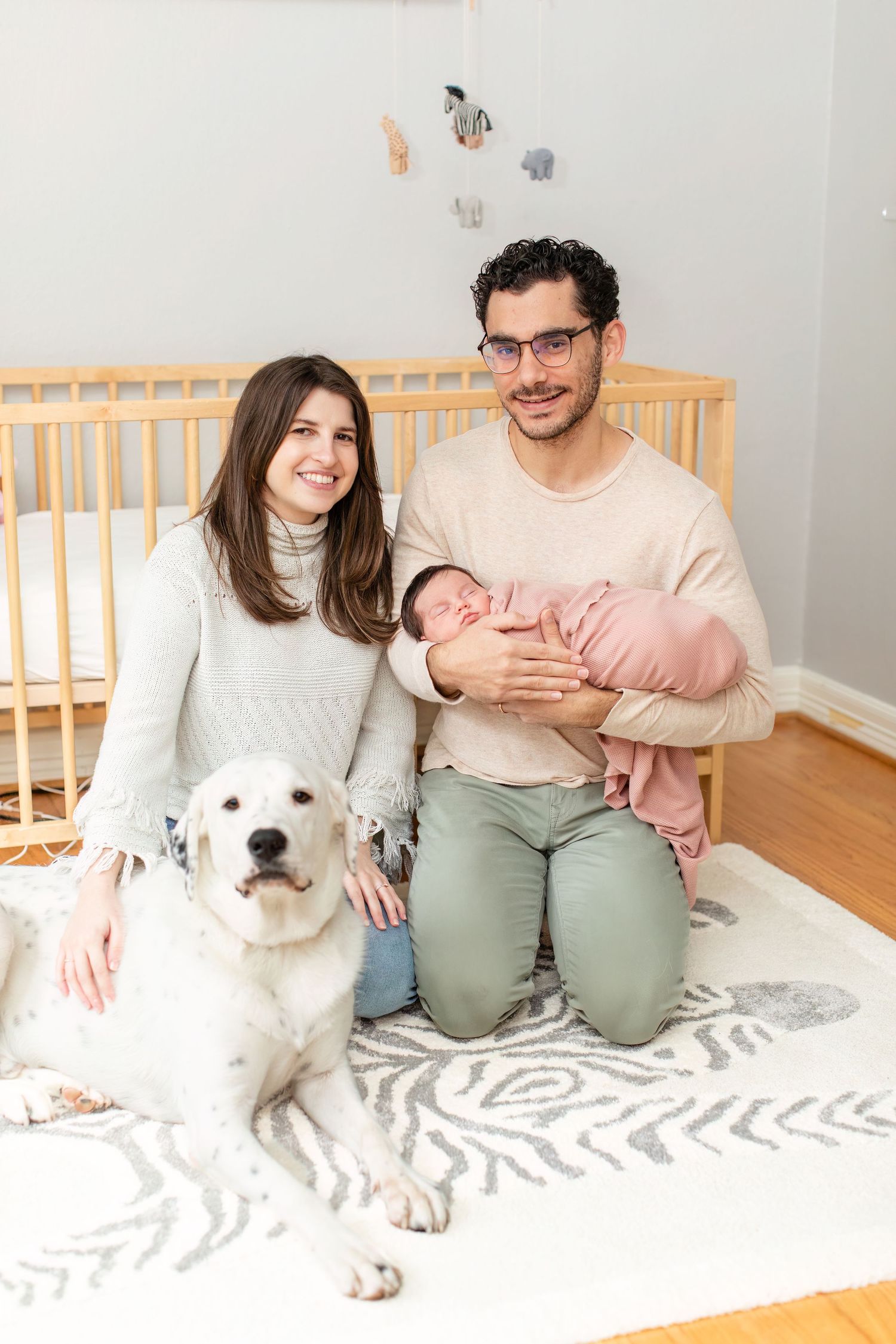 Family poses with white dog in nursery with wooden crib and mobile decorations.