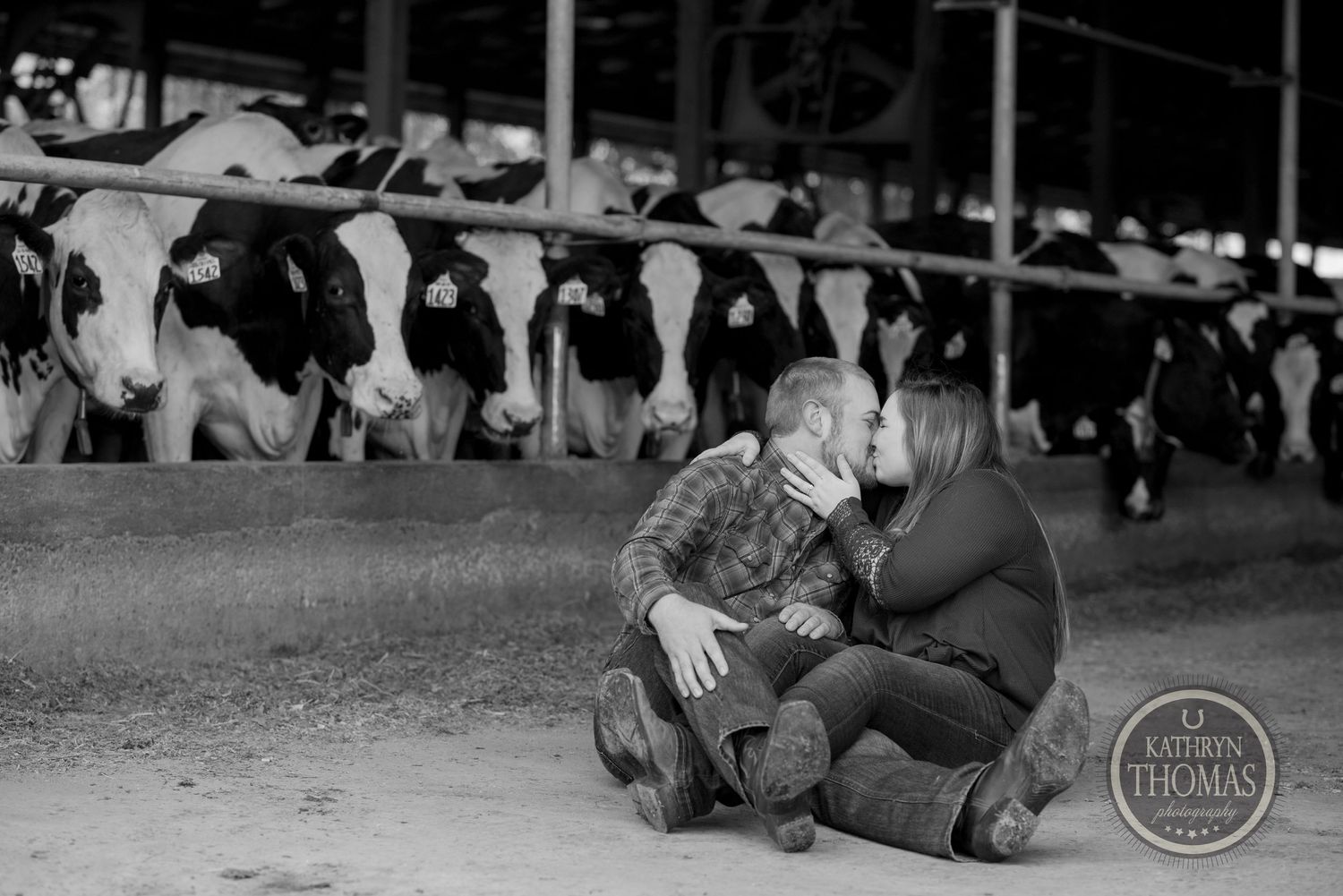 Black and white photo of couple sharing intimate moment in dairy barn setting.