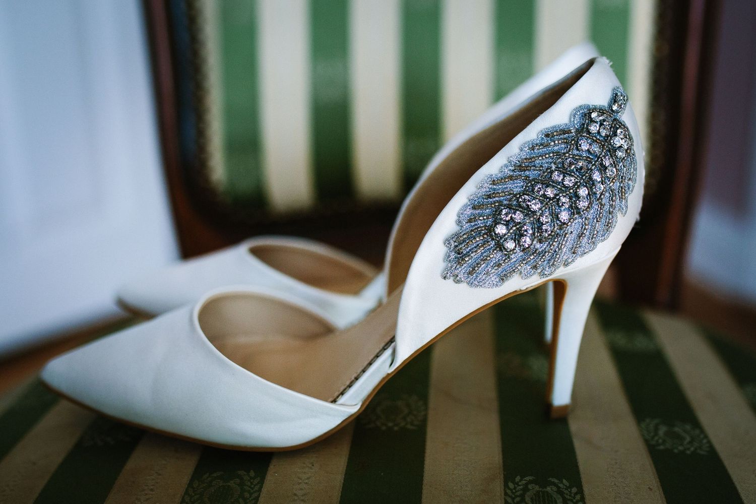Elegant white wedding shoe with sparkly crystal embellishments on a striped green chair.