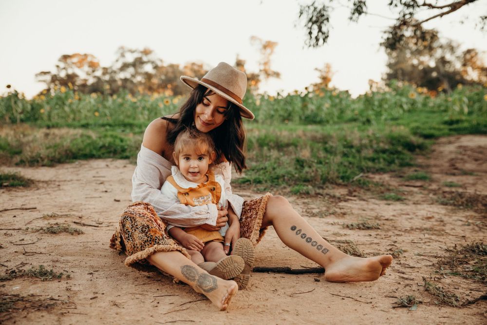 Boho mother in floral skirt and hat sitting with her child on sandy ground.