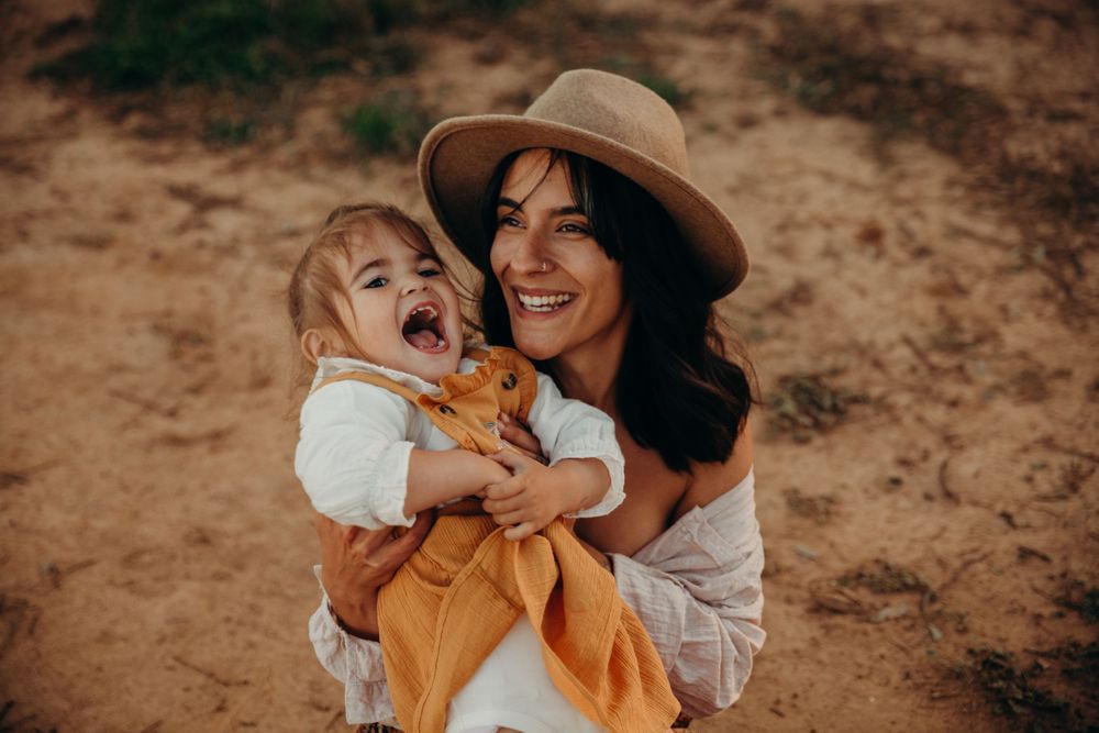 Smiling mother in straw hat laughing with happy toddler in orange dress outdoors.