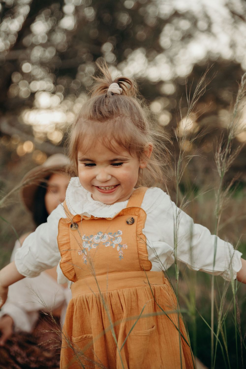 Joyful toddler girl in mustard pinafore dress playing in tall grass outdoors.