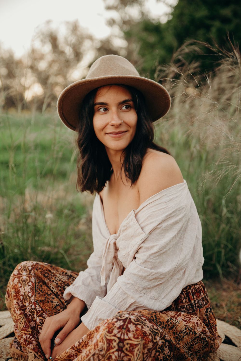 Bohemian woman in wide-brim hat and floral pants sitting outdoors in nature.