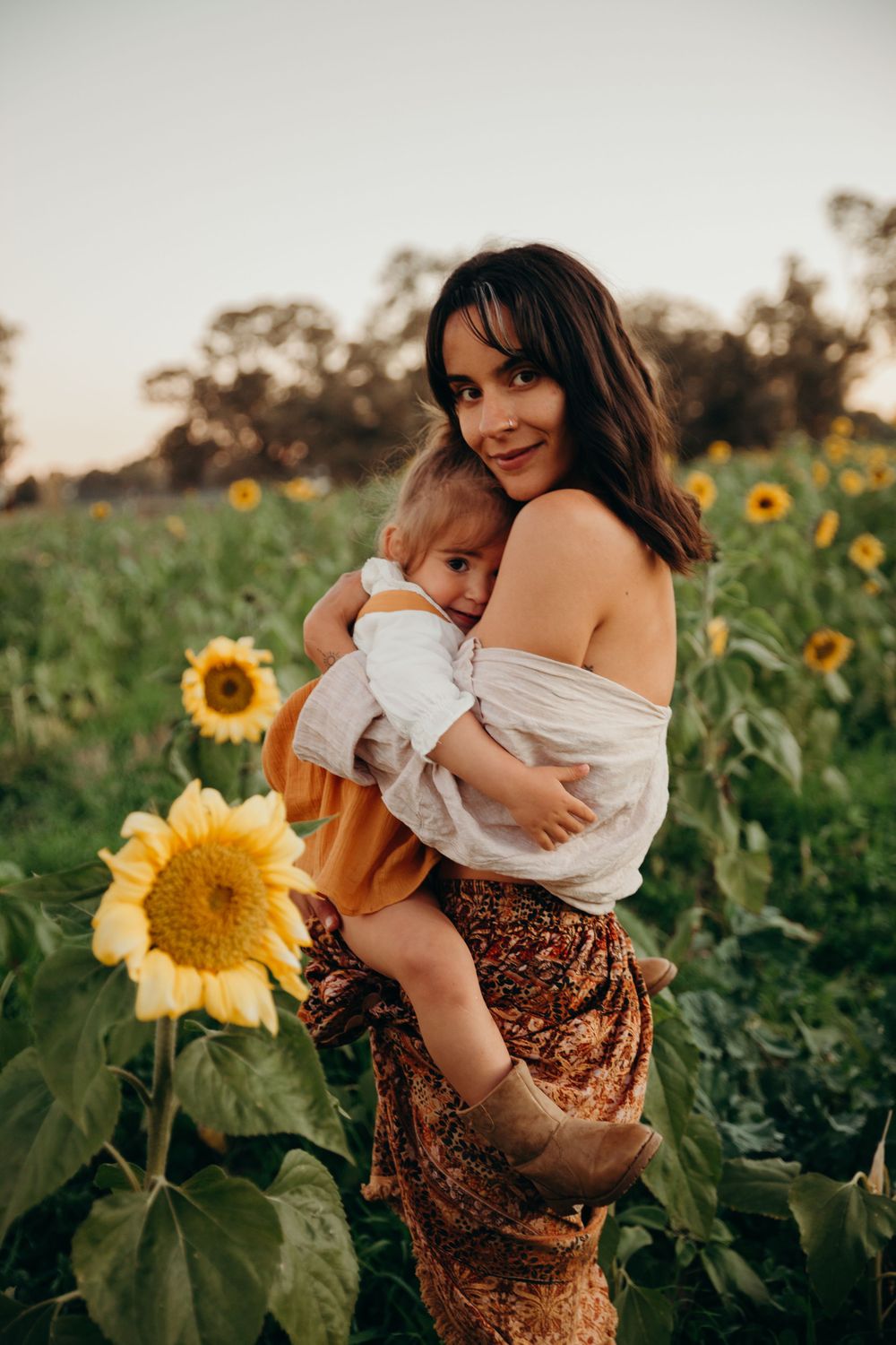 Mother holding sleeping toddler in sunflower field at golden hour sunset.