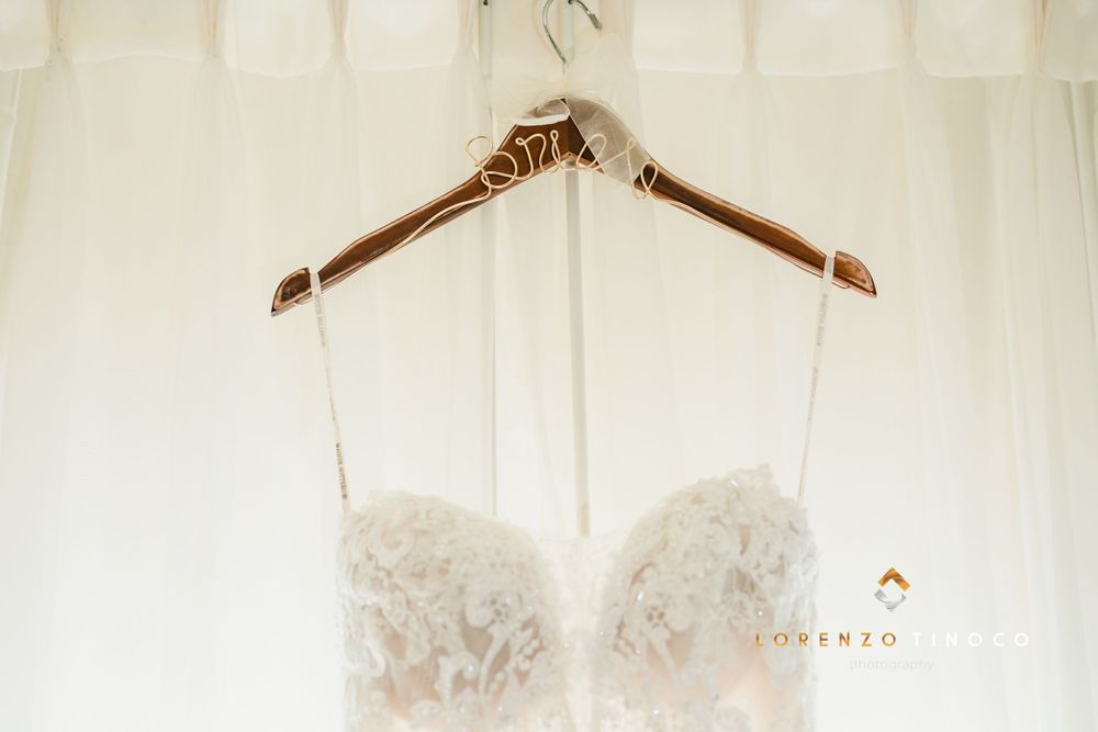 White lace wedding dress hanging on wooden hanger against sheer curtain background.