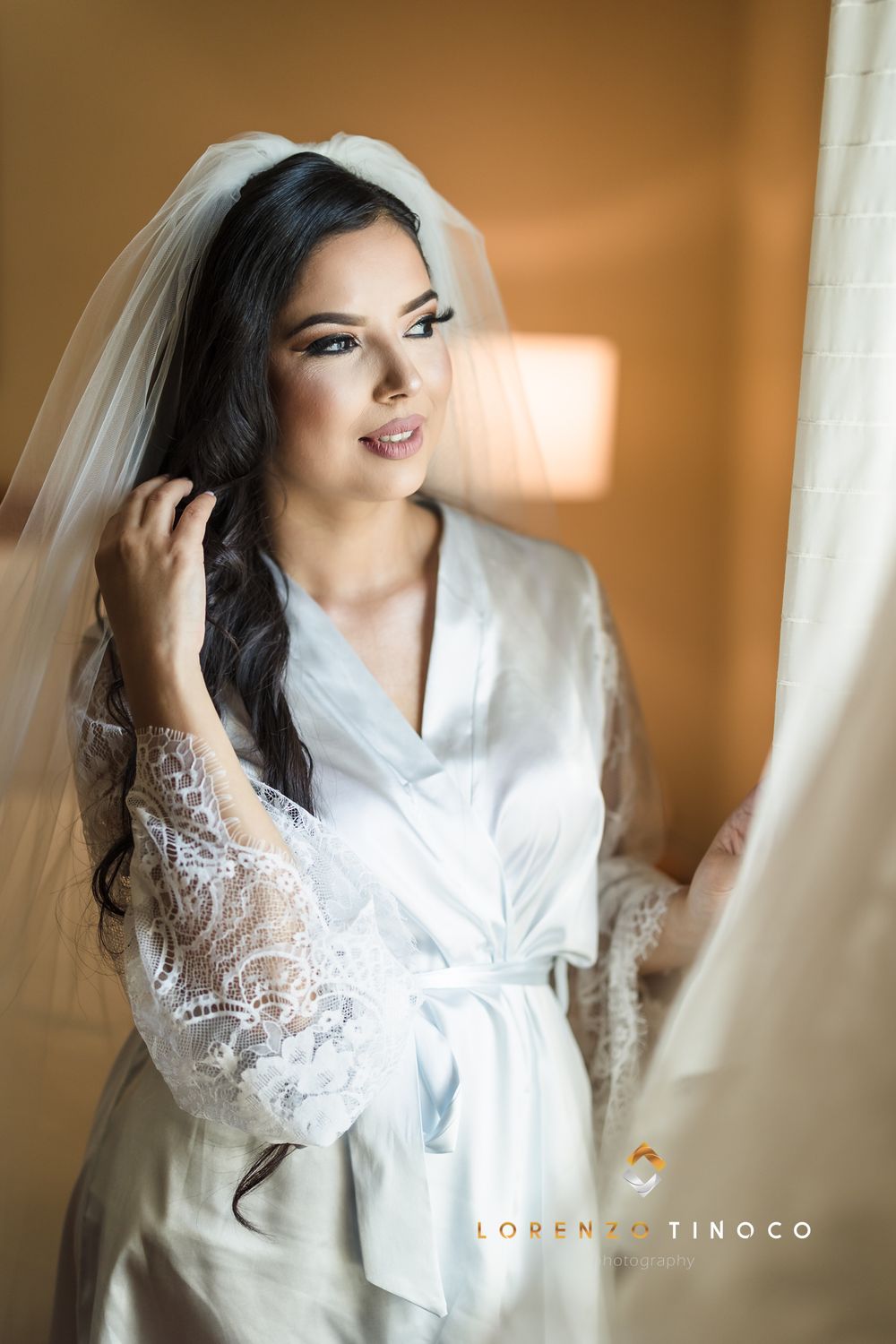 Bride in white lace robe poses with veil near window during wedding preparations.