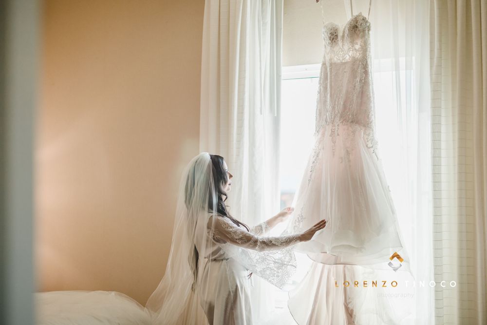 Side view of person in bridal attire admiring wedding dress hanging by window.