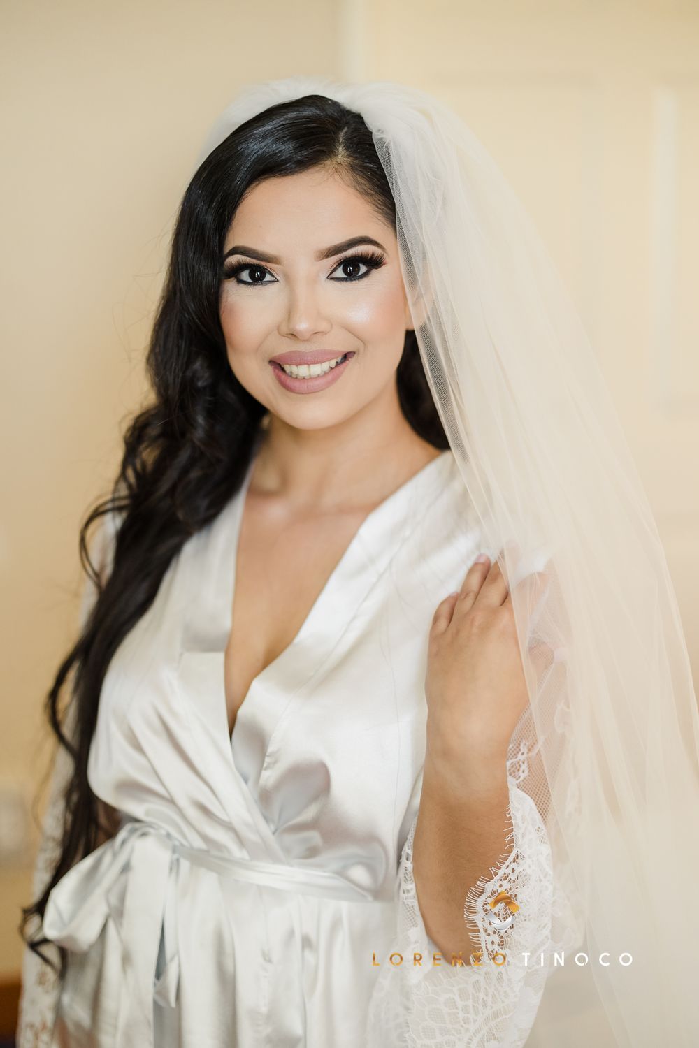 A bride wearing a white silk robe and bridal veil smiles while getting ready on her wedding day.