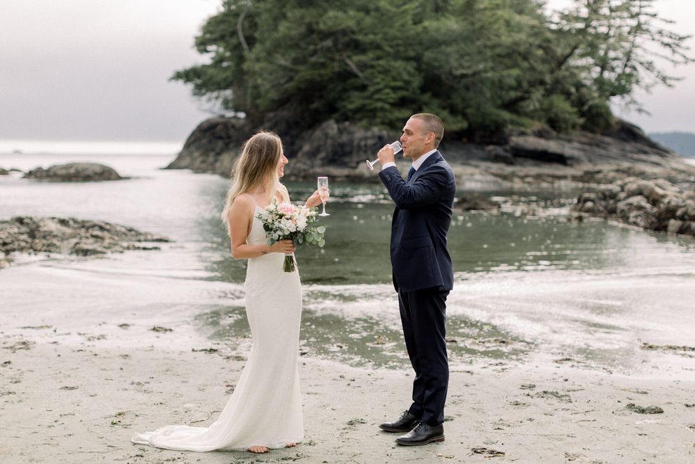 Groom photographs bride holding bouquet during beach elopement with rocky island backdrop.