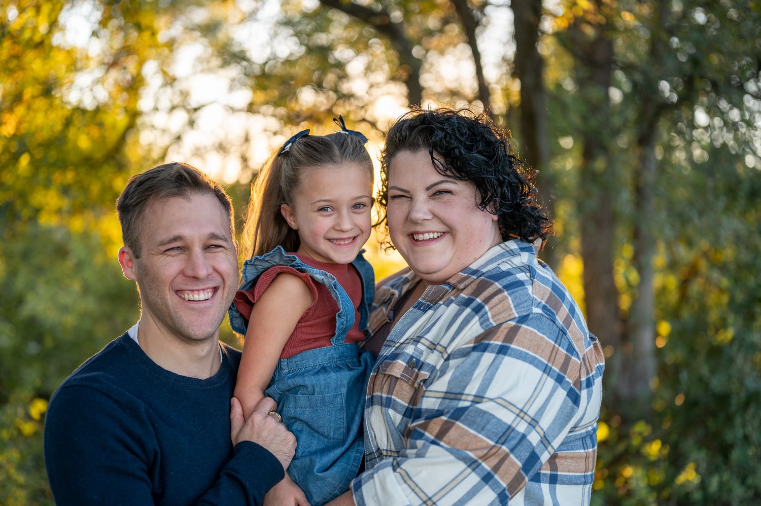 A family poses together outdoors with sunlit autumn trees in the background during golden hour.