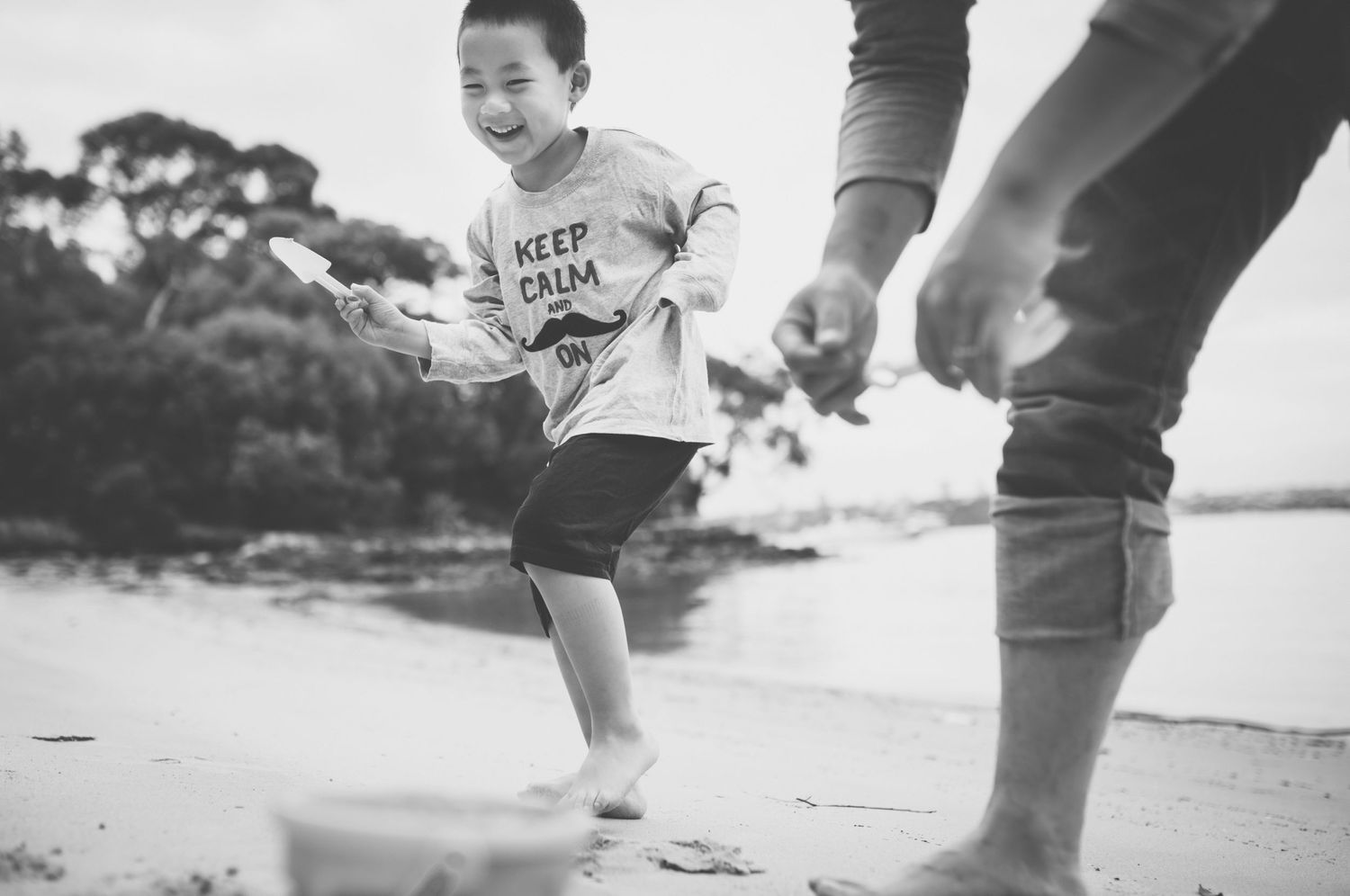 Young boy in Keep Calm shirt running barefoot on beach in black and white photo.