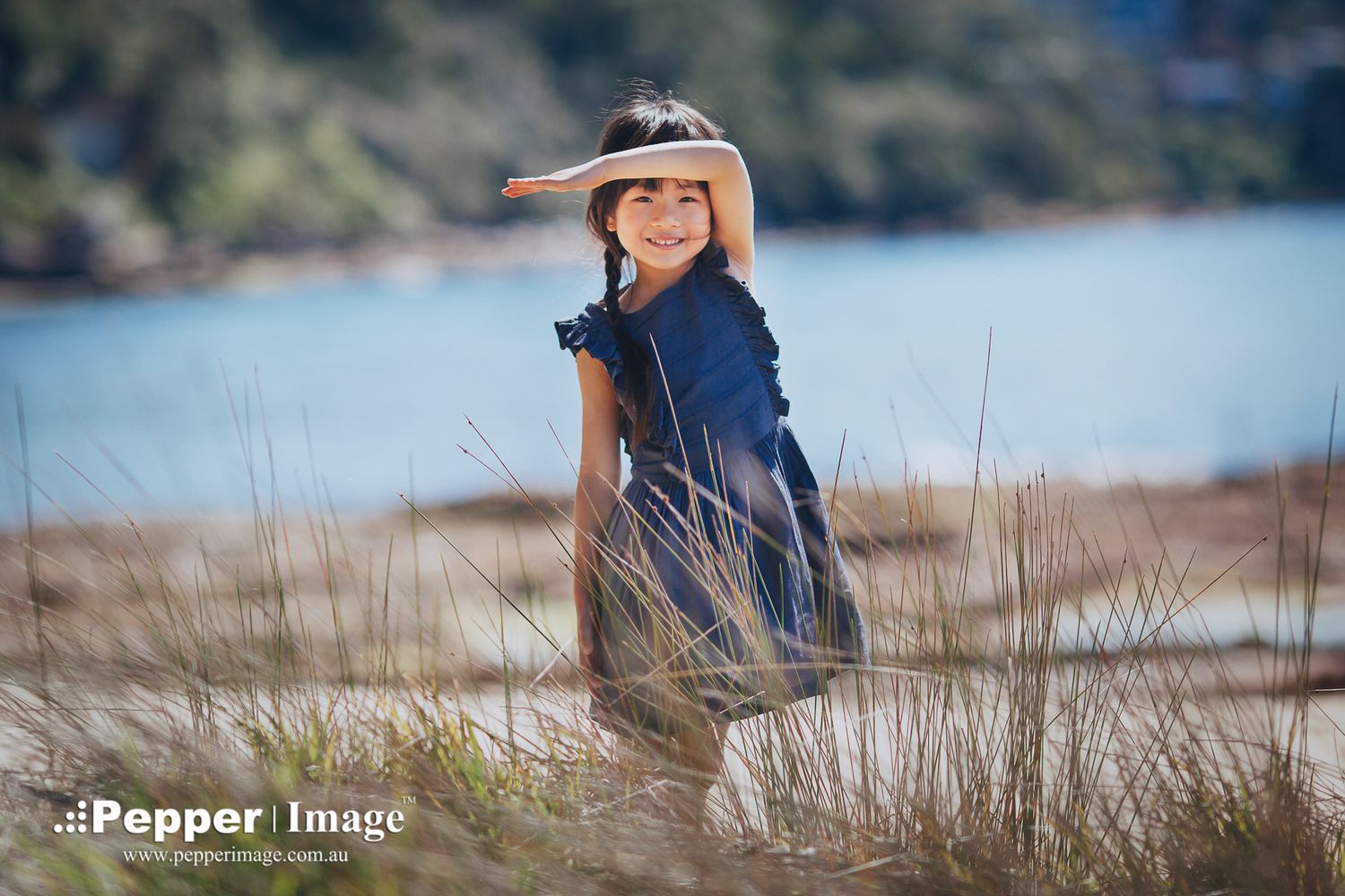 Young girl in navy dress and sun hat smiling outdoors near water in tall grass.