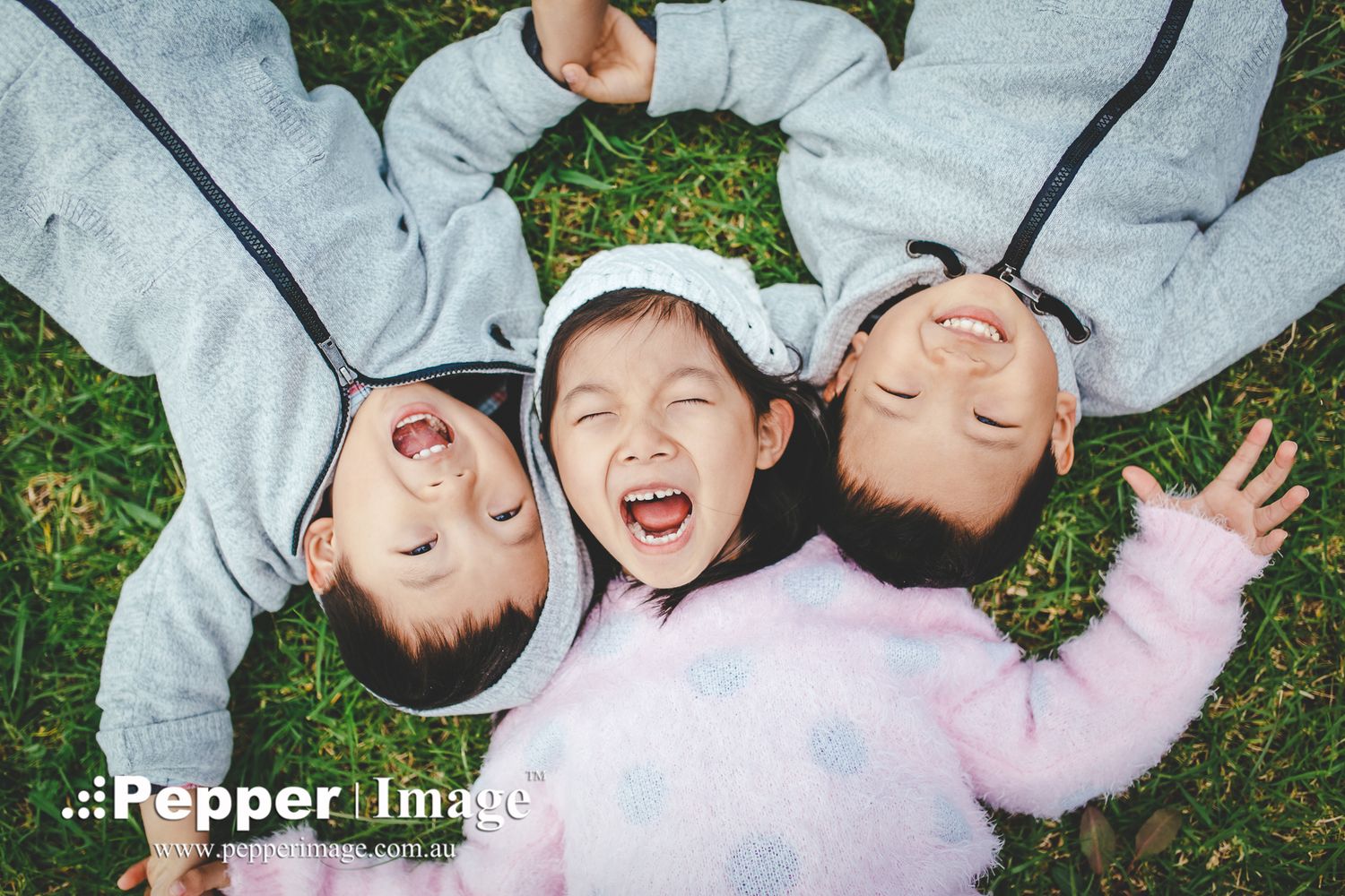 Three happy Asian children laughing while lying on green grass in a circle.