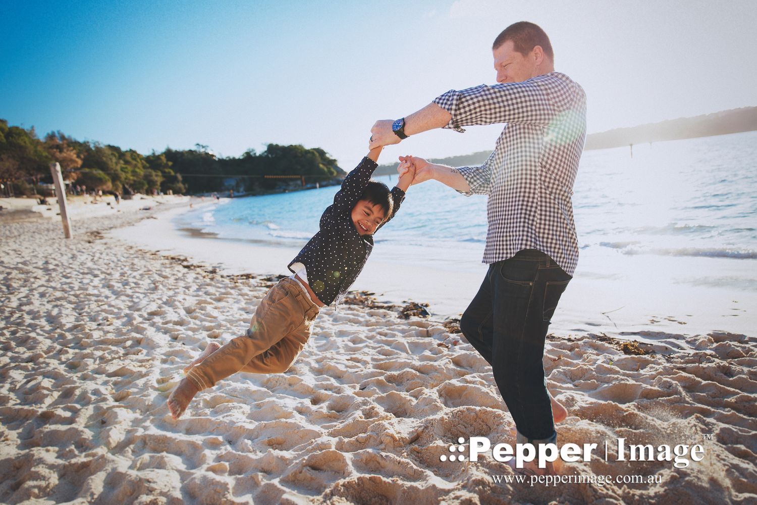 A father playfully swings his young son by the arms on a bright sunny beach.