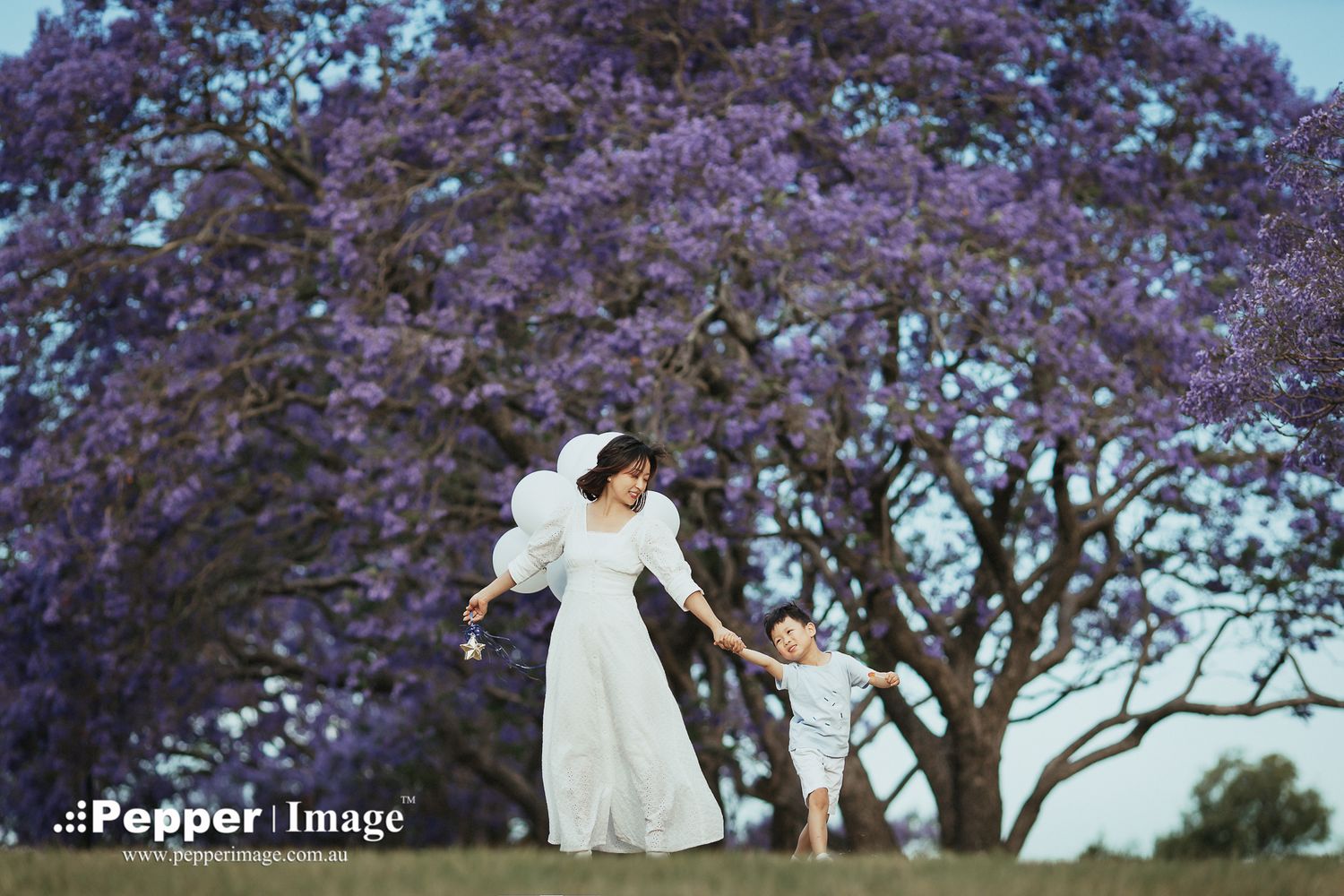 Mother in white dress dancing with child beneath a stunning purple jacaranda tree in bloom.